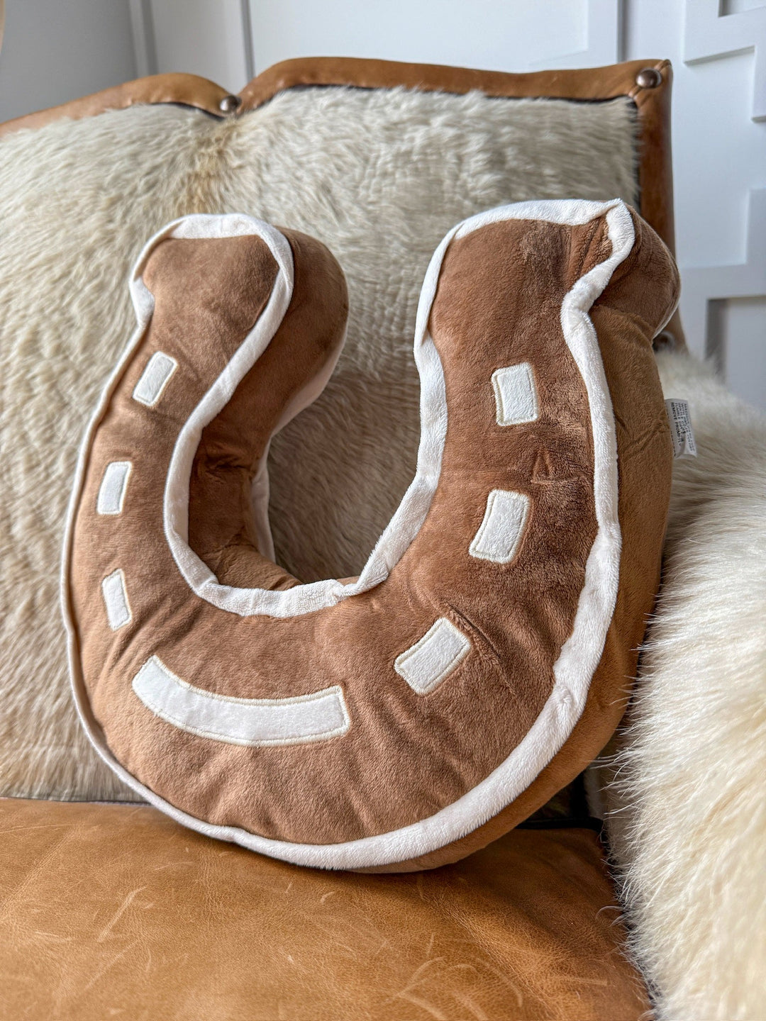 The Lucky Saddle Tan Horseshoe Pillow by Savannah Sevens western life{&}style, featuring brown and white accents, rests on a beige cushion atop a saddle tan leather chair—an ideal addition to any western bedroom.