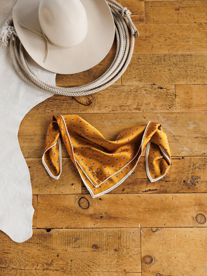 A beige cowboy hat, the Lucky You Neck Scarf by Savannah Sevens western life{&}style, coiled rope, and white cowhide are arranged on a rustic wooden floor.