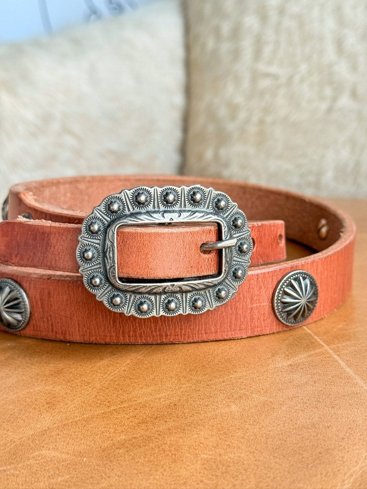 A close-up of the Luray Belt by Savannah Sevens western life{&}style, featuring cognac leather with an ornate silver buckle and matching conchos, resting on a tan surface against a soft, textured background.