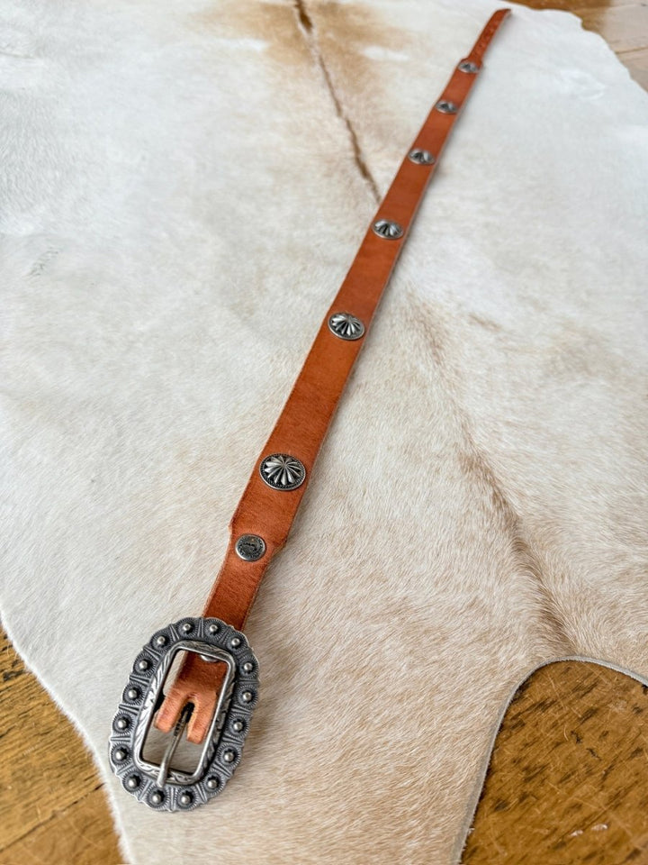 A Savannah Sevens western life{&}style Luray Belt featuring cognac leather, a decorative silver buckle, and concho accents is displayed on a light animal hide over a wooden surface.