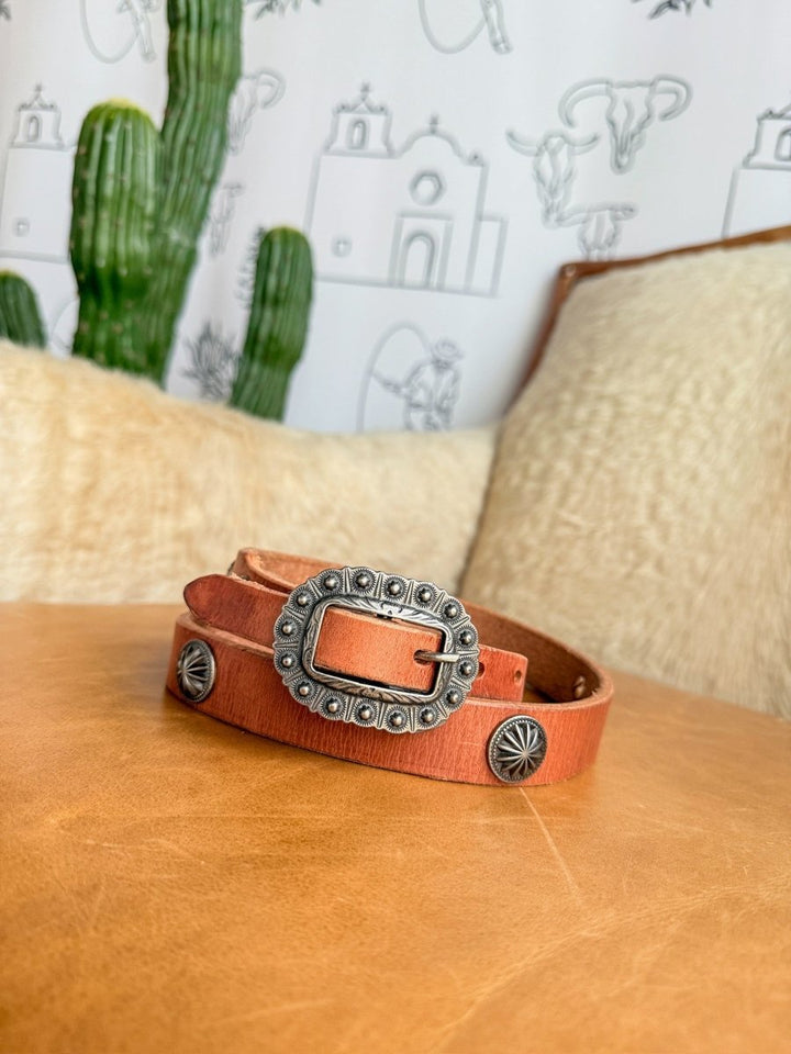 The Luray Belt by Savannah Sevens western life{&}style, featuring cognac leather and a silver buckle with concho details, rests on tan leather. A cushion, green cactus, and Southwestern-themed wallpaper appear in the background.