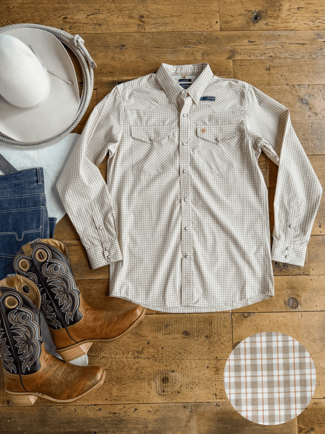 The Lyford Ariat Men’s Plaid Pearl Snap shirt from Savannah Sevens western life{&}style is laid flat on a wooden floor, accompanied by cowboy boots, blue jeans, and a white cowboy hat placed nearby. A circular inset provides a close-up view of the shirt’s plaid pattern and signature pearl snaps.
