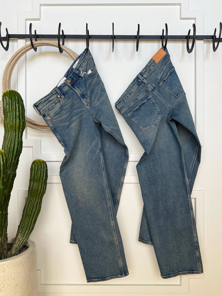 Two pairs of The Lyndon Premier Rise Relaxed Straight Leg Jeans by Savannah Sevens western life{&}style hang from hooks on a white paneled wall. A potted cactus sits nearby on wooden flooring, creating a cozy indoor vibe.