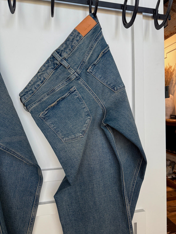 A pair of Savannah Sevens western life{&}style The Lyndon Premier Rise Relaxed Straight Leg Jeans hangs on a black hook rack against a white door, showcasing the back pockets and a brown leather patch on the waistband.