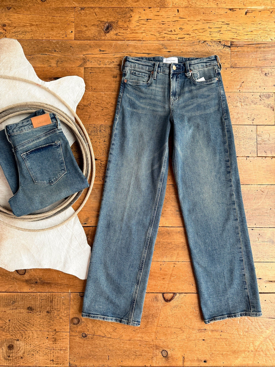 A pair of blue jeans lies flat on a wooden floor next to a folded Lyndon Premier Rise Relaxed Straight Leg Jean from Savannah Sevens western life{&}style, a coil of rope, and a white textured fabric.