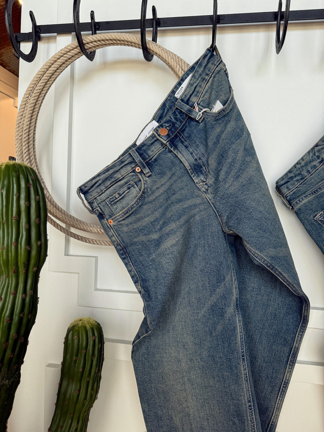 The Lyndon Premier Rise Relaxed Straight Leg Jean by Savannah Sevens western life{&}style hangs from a black metal hook rack on a white wall, with a coiled rope and two tall green cacti nearby.