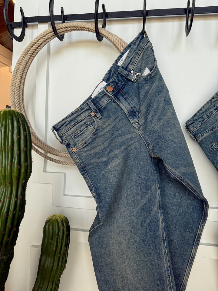 The Lyndon Premier Rise Relaxed Straight Leg Jean by Savannah Sevens western life{&}style hangs from a black metal hook rack on a white wall, with a coiled rope and two tall green cacti nearby.