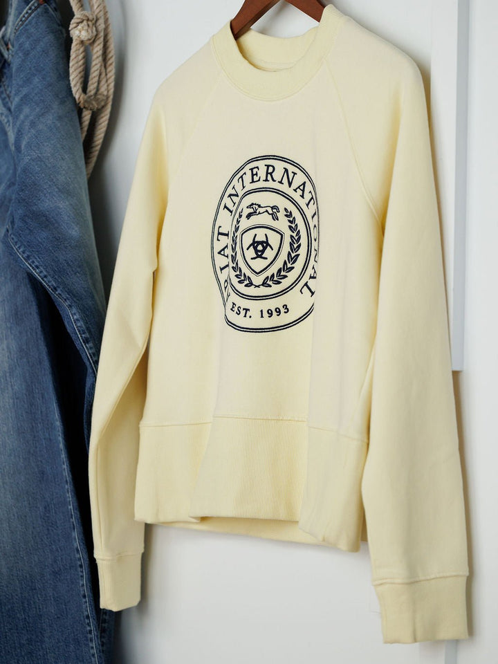The Lynwood Ariat Butter Yellow Pullover by Savannah Sevens western life{&}style, an organic cotton top featuring a black crest logo and “INTERNATIONAL EST. 1993,” hangs on a wooden hanger beside denim jeans and coiled rope.