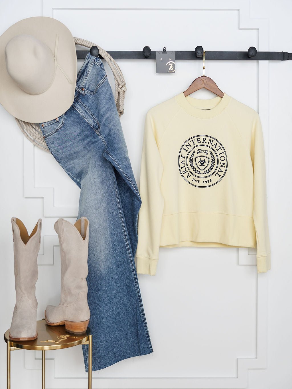 A beige cowboy hat, blue jeans, and the Lynwood Ariat Butter Yellow Pullover by Savannah Sevens western life{&}style with a circular emblem hang on a black wall rack. Light beige cowboy boots rest on a gold stool below against white paneling.