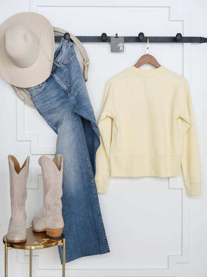 The Lynwood Ariat Butter Yellow Pullover by Savannah Sevens western life{&}style hangs alongside a beige cowboy hat and blue jeans on wall hooks, with light tan cowboy boots displayed on a small round table against a white wall.