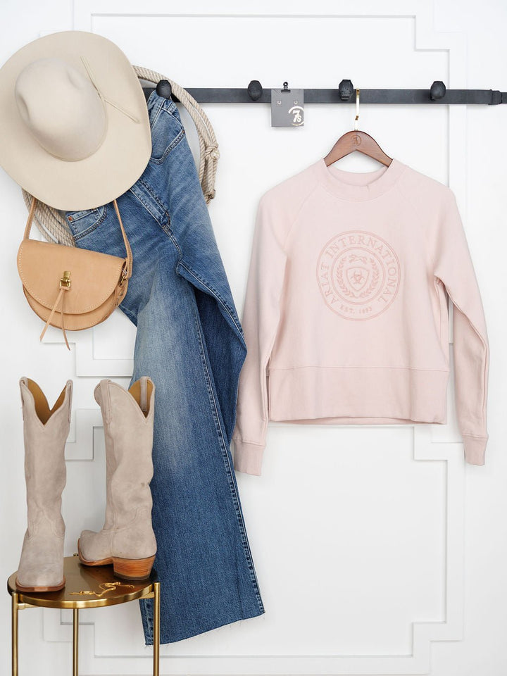 The Lynwood Ariat Light Pink Pullover by Savannah Sevens western life{&}style hangs on a hanger against a white wall, next to a beige hat, tan crossbody bag, blue jeans, and taupe boots arranged on a wall hook and stool.