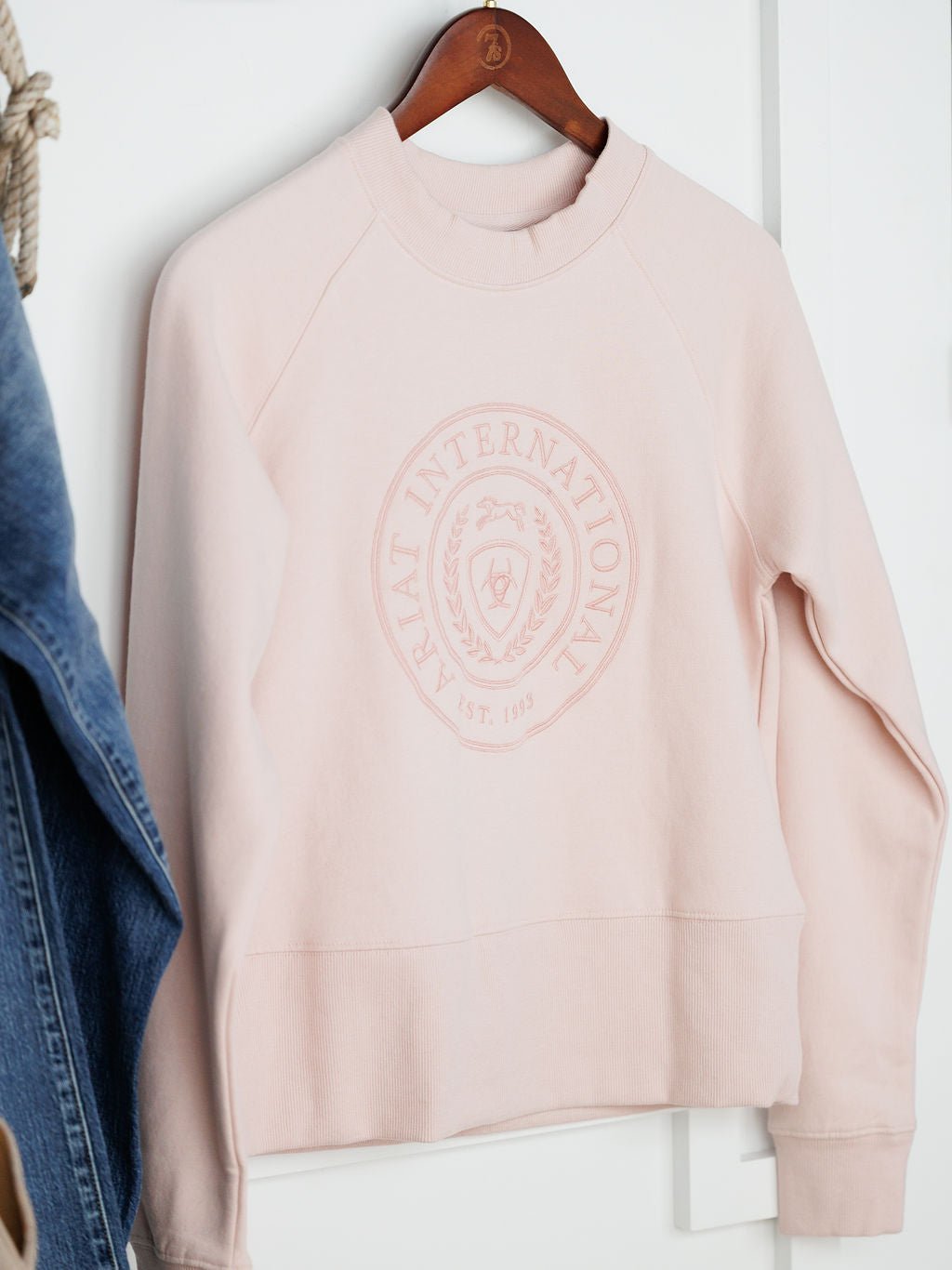 The Lynwood Ariat Light Pink Pullover by Savannah Sevens western life{&}style is a round neck, long sleeve organic cotton sweatshirt with an embossed “ARIAT INTERNATIONAL EST. 1993” emblem, displayed on a wooden hanger.