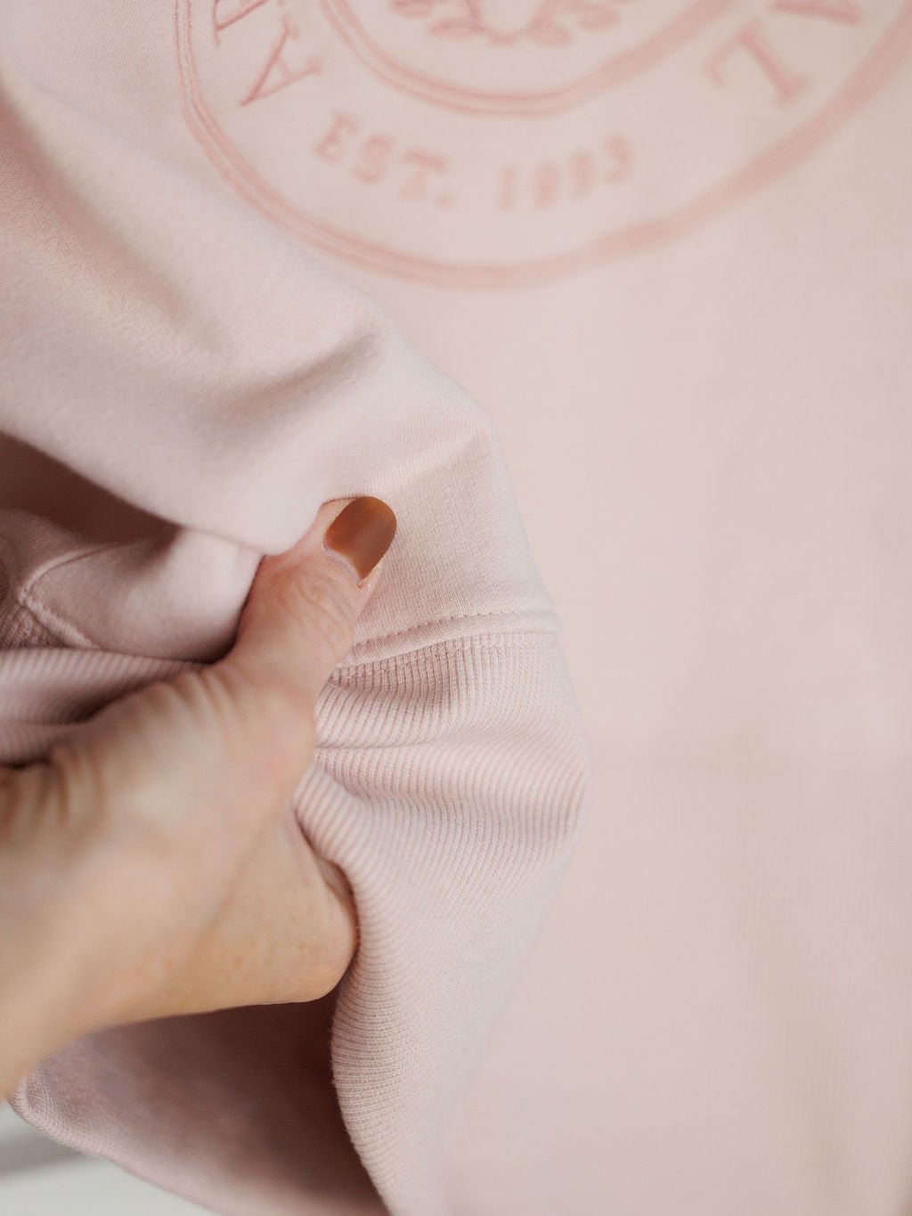 A close-up of a hand holding the sleeve of the Lynwood Ariat Light Pink Pullover by Savannah Sevens western life{&}style, showing part of a circular logo and text on the soft organic cotton fabric.