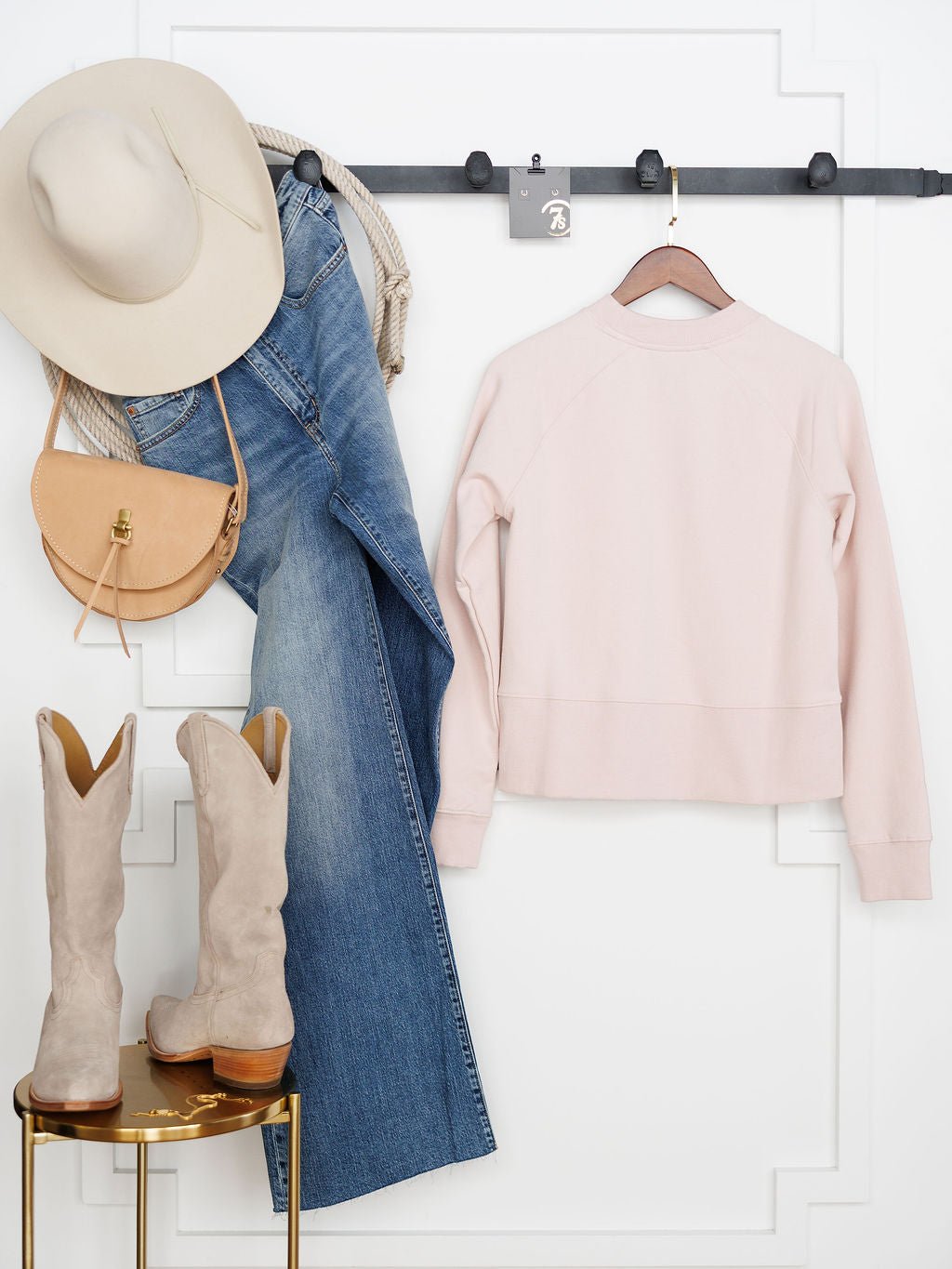 A beige wide-brim hat, blue jeans, a tan purse, and the Lynwood Ariat Light Pink Pullover by Savannah Sevens western life{&}style hang on a wall rack. Below, fleece-lined beige cowboy boots sit on a small round table.