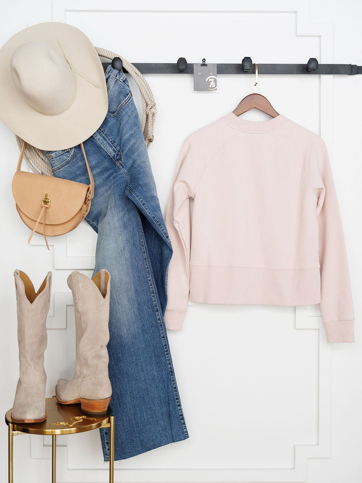A beige wide-brim hat, blue jeans, a tan purse, and the Lynwood Ariat Light Pink Pullover by Savannah Sevens western life{&}style hang on a wall rack. Below, fleece-lined beige cowboy boots sit on a small round table.