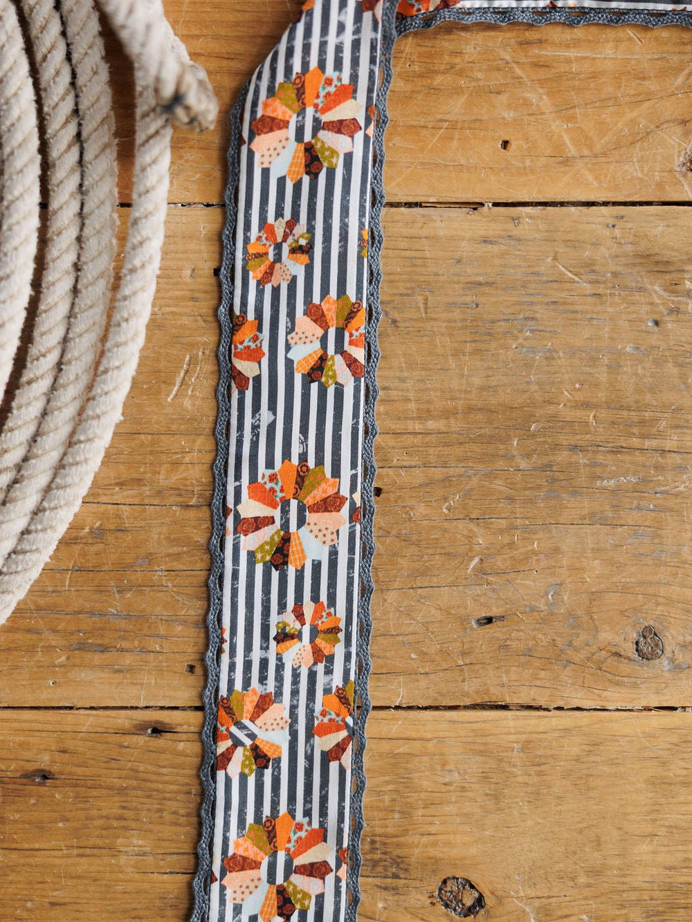 The Mabel Twilly by Savannah Sevens western life{&}style, featuring orange, red, and yellow floral stripes on satin, rests on a wooden surface beside a coiled white rope.