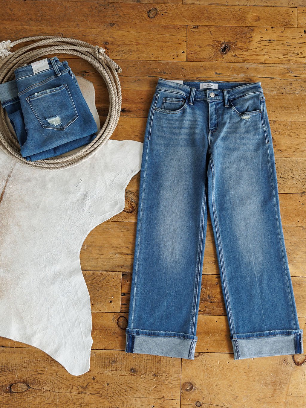 The Mack High Rise Relaxed Straight Leg Jean by Savannah Sevens western life{&}style is laid flat on wood next to folded Vervet jeans on a white cowhide rug and a coiled rope, adding a rustic, western touch.