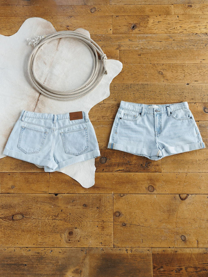 Two pairs of The Mackie Shorts by Savannah Sevens western life{&}style are laid flat on a wooden floor—one showing the front, one the back. A looped rope sits on a white animal hide rug in the upper left corner.