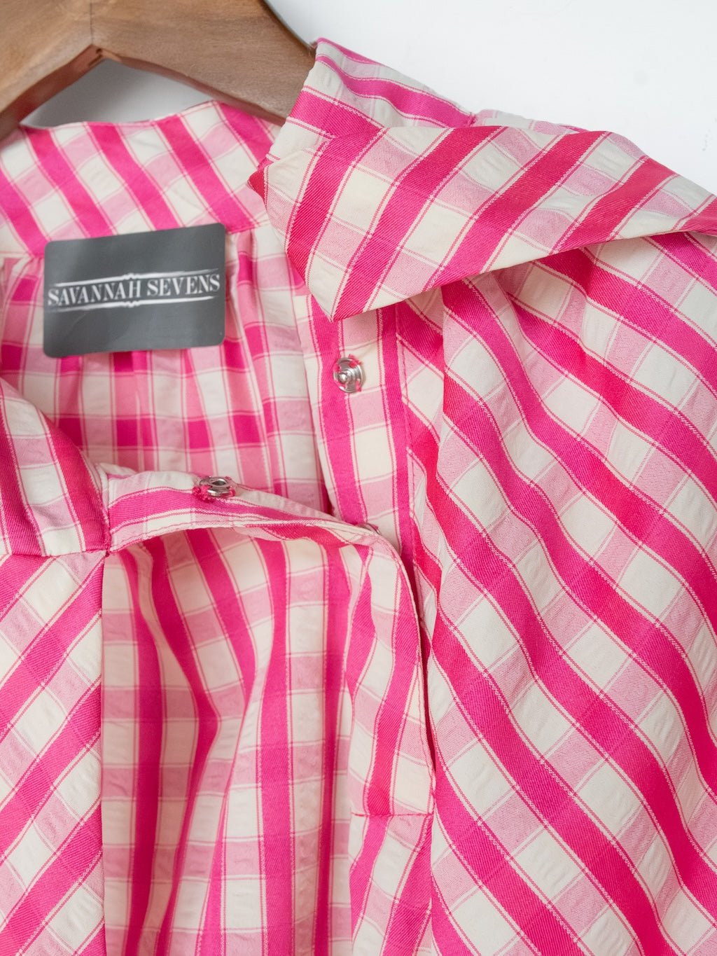 A close-up of the Madelia Pink Gingham Puff Sleeve Top by Savannah Sevens western life{&}style, with diagonal stripes, hanging on a wooden hanger.