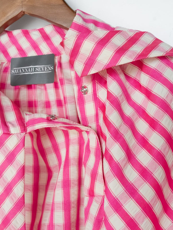 A close-up of the Madelia Pink Gingham Puff Sleeve Top by Savannah Sevens western life{&}style, with diagonal stripes, hanging on a wooden hanger.