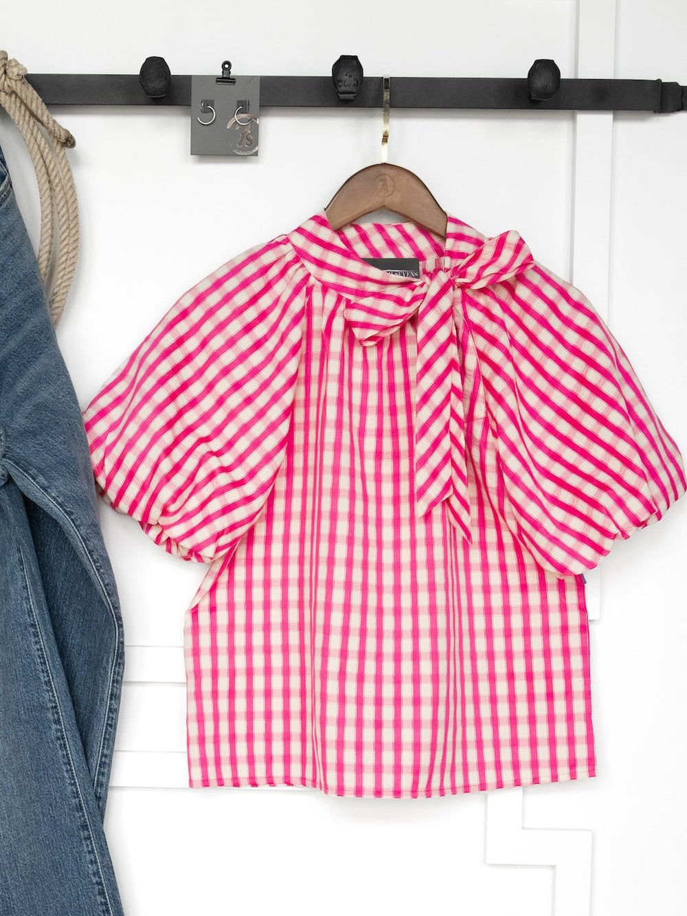 The Madelia Pink Gingham Puff Sleeve Top by Savannah Sevens western life{&}style, featuring a large bow collar, hangs on a wooden hanger beside blue jeans on a wall rack.