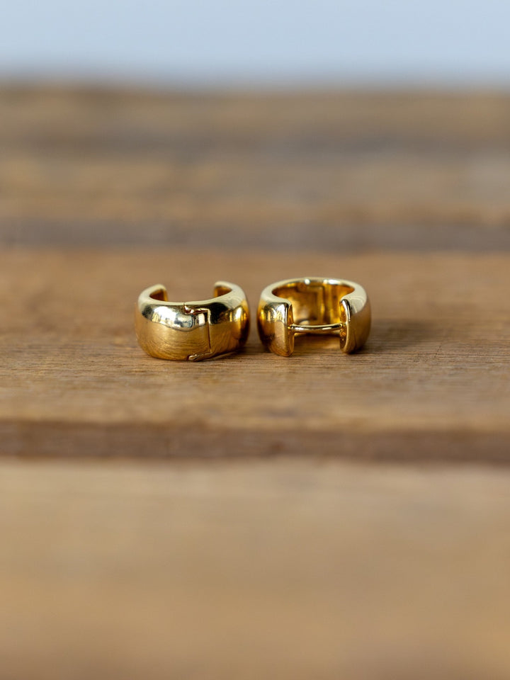 The Madera Gold Mini Hoop Earrings by Savannah Sevens western life{&}style are shown on a wooden surface—one upright, the other on its side, highlighting the hinge and clasp design.