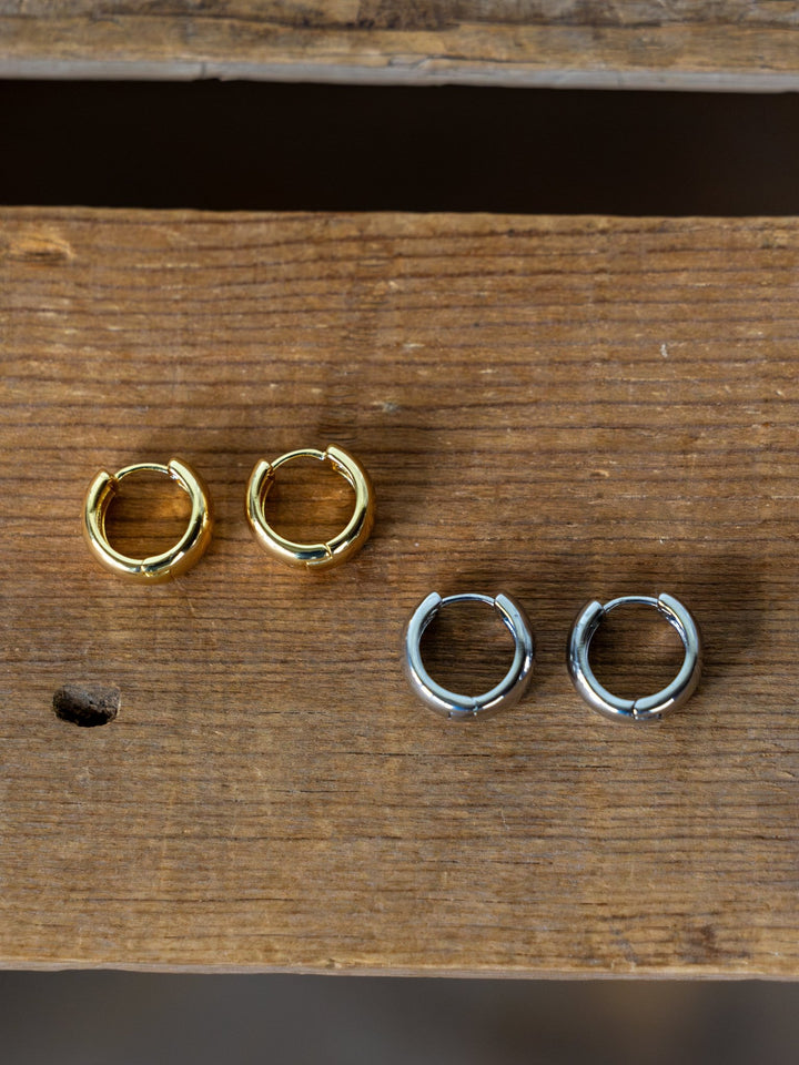 The Savannah Sevens western life{&}style Madera Gold/Silver Mini Hoop Earrings—one gold, one silver—are displayed side by side on a wooden surface.