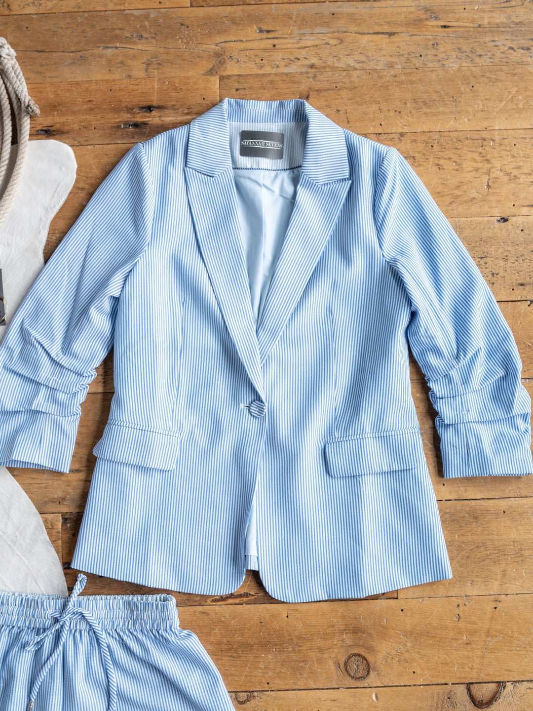 The Malin {Set} by Savannah Sevens western life{&}style, a blue and white pinstriped single-button blazer paired with matching high-waisted shorts, is displayed flat on a wooden floor beside a coiled rope.