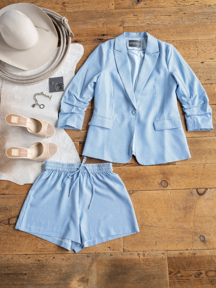 A flat lay on a wooden floor features The Malin {Set} by Savannah Sevens western life{&}style—blue and white pinstripe blazer with matching high waisted shorts—styled with beige heels, a wide-brim hat, and a key charm bracelet on a cowhide rug.