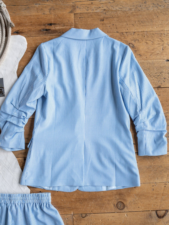 The Malin {Set} by Savannah Sevens western life{&}style features a light blue and white vertically striped long-sleeve shirt laid flat, back up, on wood, with part of the matching high-waisted shorts from the pinstriped set visible at the edge.