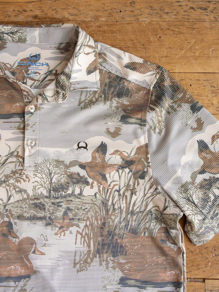 The Mallard Cinch Men's Graphic Polo by Savannah Sevens western life{&}style is a short-sleeve collared shirt featuring a brown, tan, and green nature-inspired duck hunting print, displayed on a wooden surface. This polo offers moisture-wicking fabric, a three-button placket, and an embroidered chest logo.