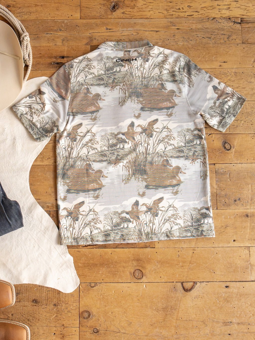The Mallard Cinch Men's Graphic Polo by Savannah Sevens western life{&}style is a short-sleeve ArenaFlex shirt with a nature-inspired print showcasing ducks, reeds, and water. Shown laid flat on a wooden floor beside a white rug, tan chair, and woven basket, this polo provides moisture wicking comfort and UV defense for all-day wear.