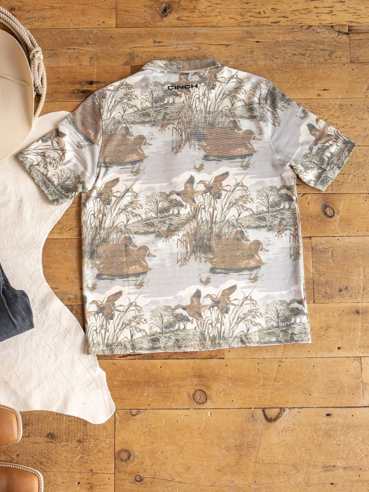 The Mallard Cinch Men's Graphic Polo by Savannah Sevens western life{&}style is a short-sleeve ArenaFlex shirt with a nature-inspired print showcasing ducks, reeds, and water. Shown laid flat on a wooden floor beside a white rug, tan chair, and woven basket, this polo provides moisture wicking comfort and UV defense for all-day wear.