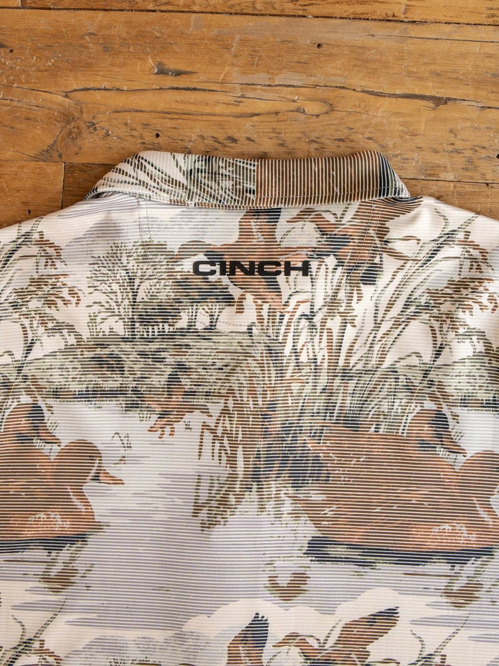 A close-up of the Savannah Sevens western life{&}style Mallard Cinch Men's Graphic Polo showcases a nature-inspired print with ducks and reeds, a striped collar, and CINCH embroidered on the back. Designed for comfort outdoors, this polo features UV defense and moisture-wicking properties. The shirt is displayed on a wooden surface.