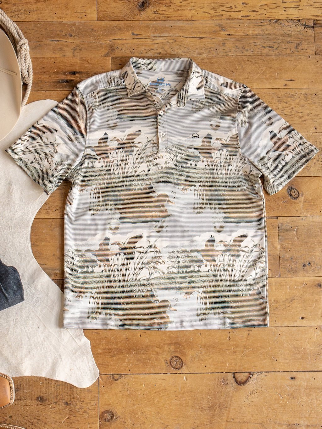The Savannah Sevens western life{&}style Mallard Cinch Men's Graphic Polo, featuring a short-sleeve collared design with an earthy nature and duck hunting scene print, is laid flat on a wooden floor beside a piece of white hide and a tan hat. This polo offers moisture-wicking comfort and UV defense, making it perfect for outdoor adventures.