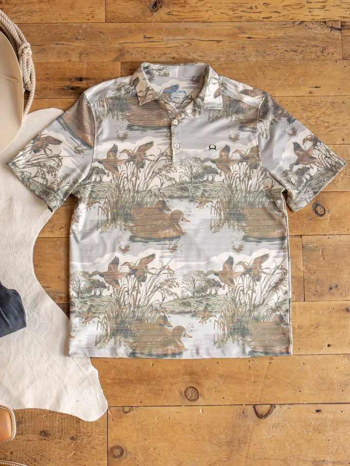 The Savannah Sevens western life{&}style Mallard Cinch Men's Graphic Polo, featuring a short-sleeve collared design with an earthy nature and duck hunting scene print, is laid flat on a wooden floor beside a piece of white hide and a tan hat. This polo offers moisture-wicking comfort and UV defense, making it perfect for outdoor adventures.