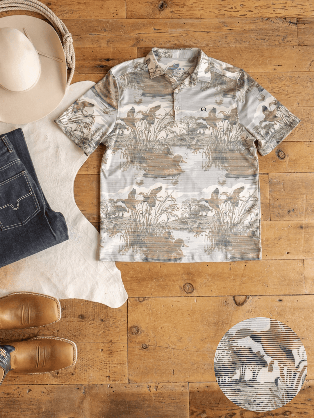 The Mallard Cinch Men's Graphic Polo by Savannah Sevens western life{&}style, a short-sleeve button-up shirt featuring a nature-inspired print, is laid on a wooden floor alongside jeans, brown cowboy boots, a beige cowboy hat, and a white cowhide rug. Crafted from moisture-wicking fabric for comfort and offering UV defense, the shirt is also pictured with a small zoomed-in detail of its distinctive print.