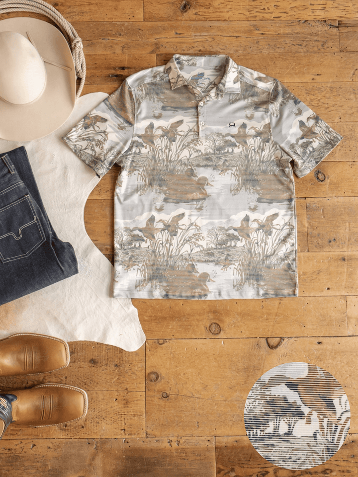 The Mallard Cinch Men's Graphic Polo by Savannah Sevens western life{&}style, a short-sleeve button-up shirt featuring a nature-inspired print, is laid on a wooden floor alongside jeans, brown cowboy boots, a beige cowboy hat, and a white cowhide rug. Crafted from moisture-wicking fabric for comfort and offering UV defense, the shirt is also pictured with a small zoomed-in detail of its distinctive print.