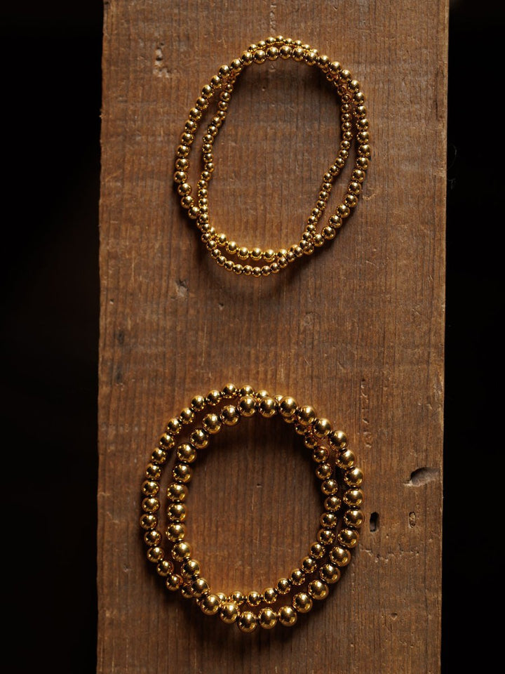 The Mancos Bracelet Set by Savannah Sevens western life{&}style includes two 18K gold plated stretch bracelets, displayed on a warm brown, rough-textured vertical wooden plank with one above the other.
