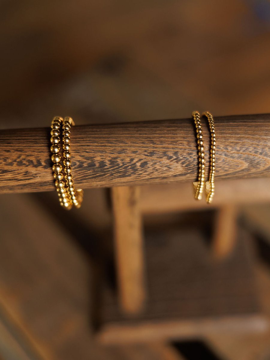 The Mancos Bracelet Set by Savannah Sevens western life{&}style features two gold bead stretch bracelets—one with large beads and one with smaller, tightly spaced 18K gold plated beads—displayed on a wooden rod against a blurred background.