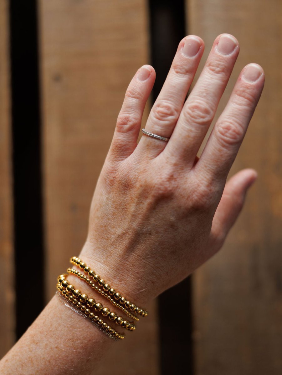 A hand with fair skin wears a thin silver ring and several Mancos Bracelet Sets by Savannah Sevens western life{&}style, featuring 18K gold plated stretch beaded bracelets, against a wooden background.