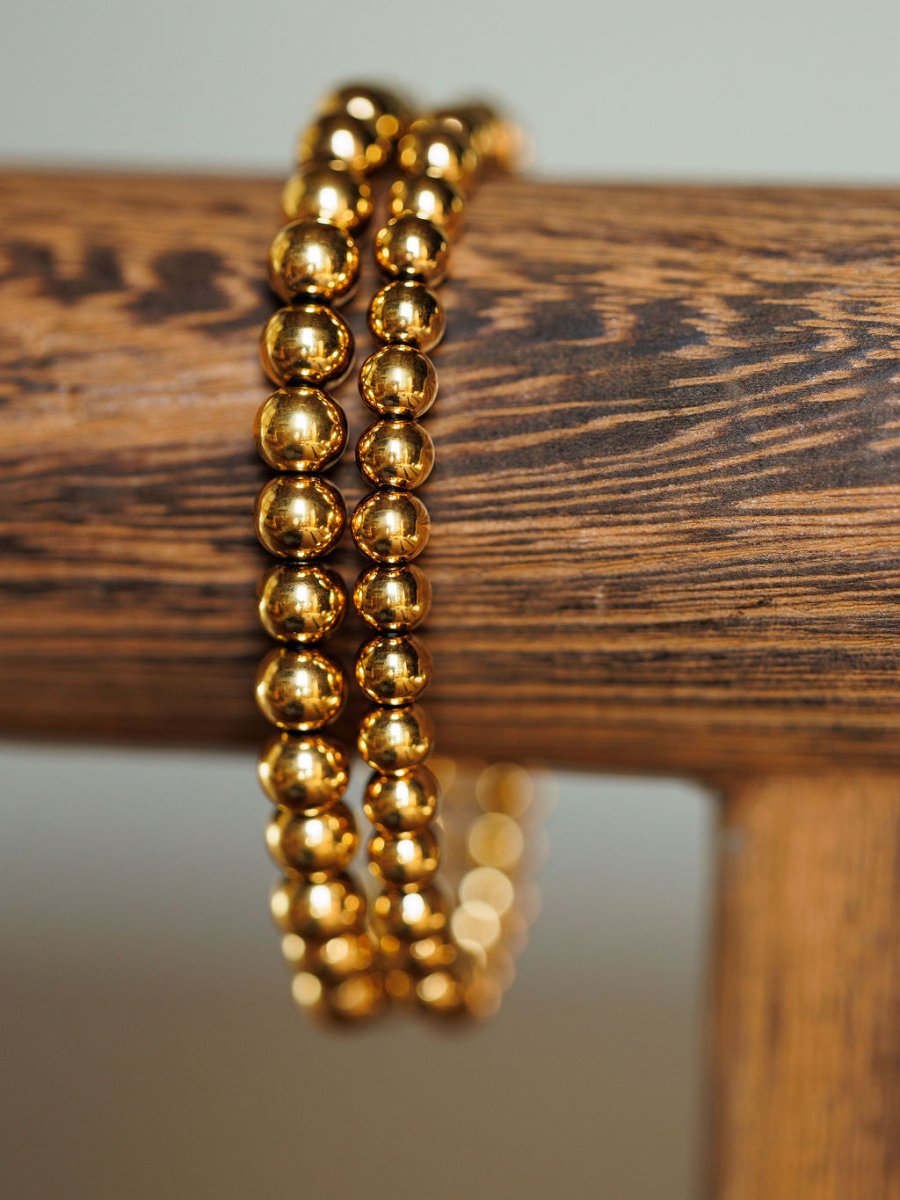 Close-up of the Mancos Bracelet Set by Savannah Sevens western life{&}style—two 18K gold plated beaded bracelets wrapped around a wooden rod, wood grain in focus, with a softly blurred background.