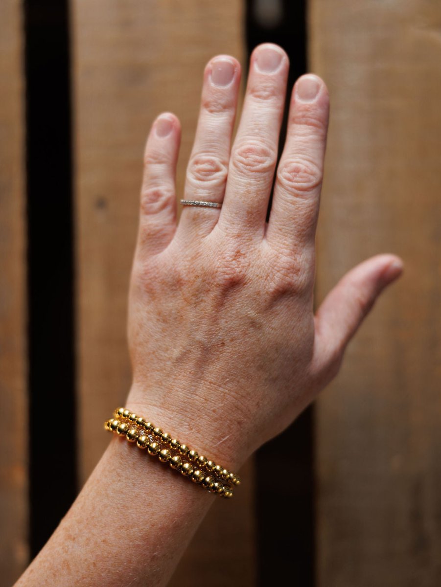 A hand wears a thin silver ring on the ring finger and the Mancos Bracelet Set by Savannah Sevens western life&style, featuring 18K gold plated double-strand beaded stretch bracelets, shown against a wooden background.