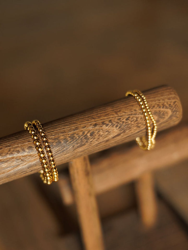 The Mancos Bracelet {Set} by Savannah Sevens western life{&}style, two 18K gold plated stretch bracelets, rests on a wooden chair. The softly blurred background highlights their shine and the wood's texture.