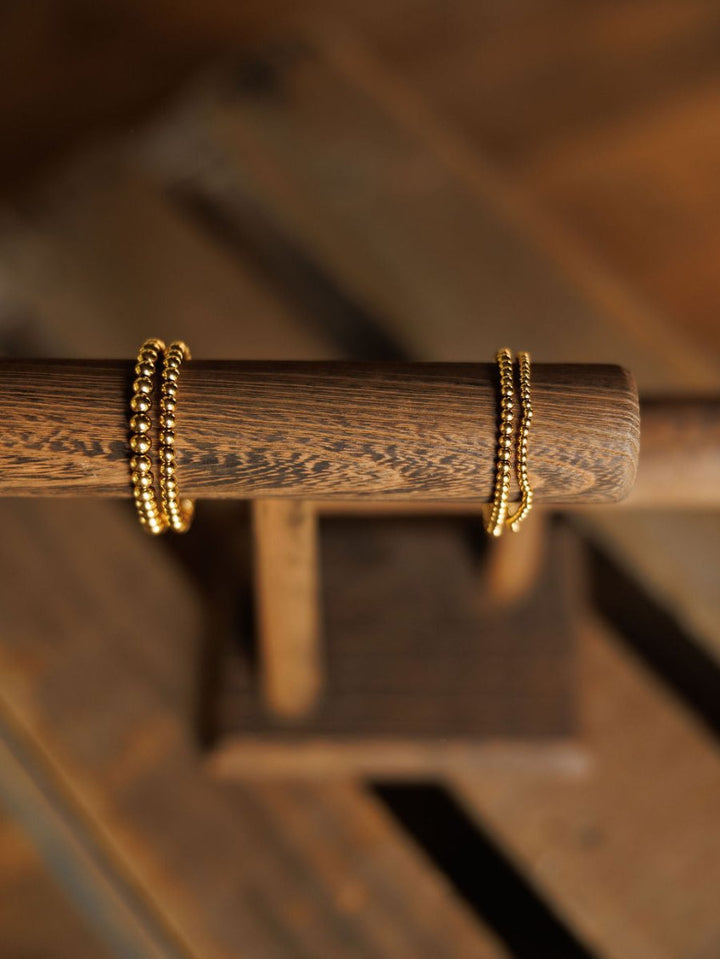 The Mancos Bracelet Set by Savannah Sevens western life{&}style features two 18K gold plated stretch bracelets displayed over a wooden rod with a blurred wood background.