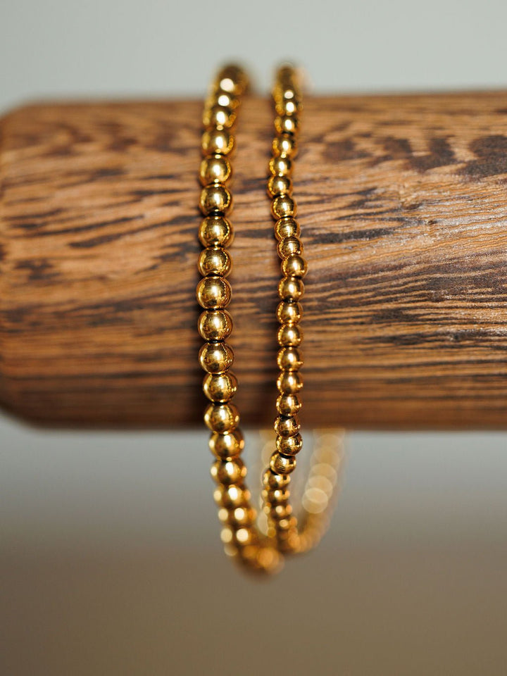 A close-up of the Mancos Bracelet Set by Savannah Sevens western life{&}style, featuring two 18K gold plated beaded stretch bracelets draped over a textured wooden rod with a softly blurred neutral background.