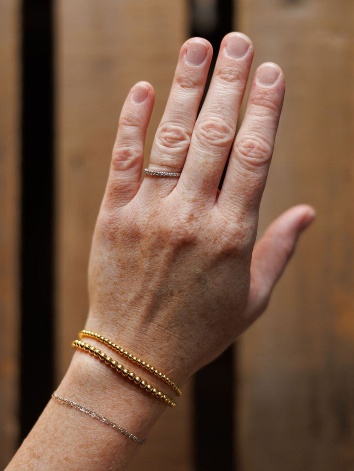 A hand wears the Mancos Bracelet Set by Savannah Sevens western life{&}style—thin silver and 18K gold plated stretch bracelets—against a blurred wooden background.