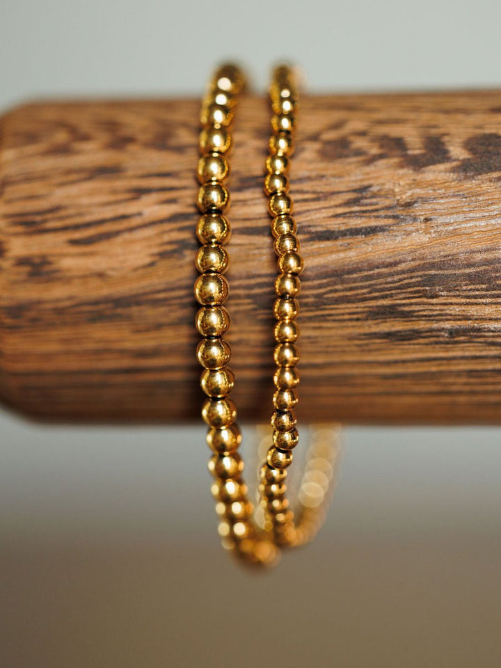 A close-up of the Mancos Bracelet Set by Savannah Sevens western life{&}style, featuring two 18K gold plated beaded stretch bracelets draped over a textured wooden rod with a softly blurred neutral background.