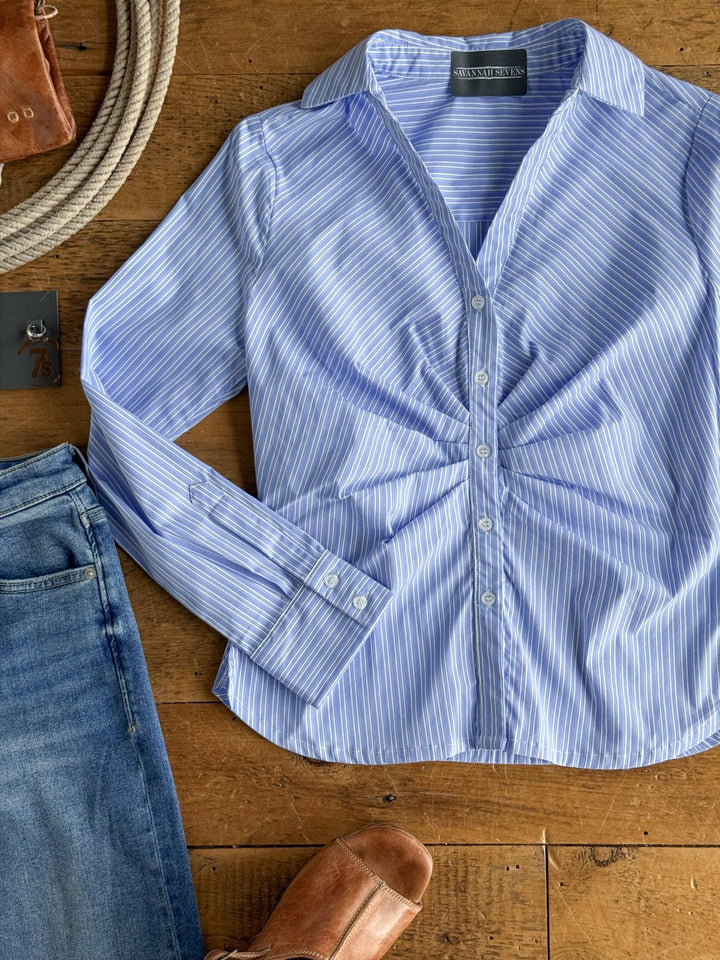 The Mandeville Blue Striped Pleat Top by Savannah Sevens western life{&}style is laid out on a wooden surface, surrounded by blue jeans, brown shoes, a coiled rope, and a partially visible leather bag.
