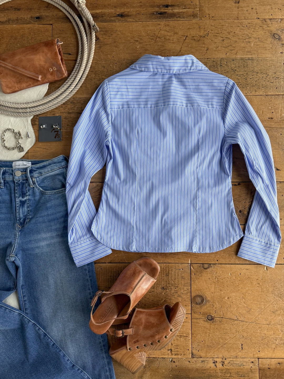 The Mandeville Blue Striped Pleat Top by Savannah Sevens western life{&}style, blue jeans, brown heeled sandals, a brown wallet, jewelry, and a coiled rope are arranged on a wooden floor.